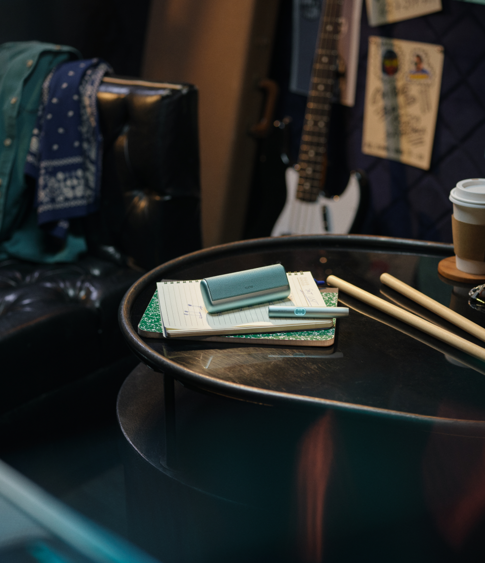 IQOS ILUMA sobre mesa con accesorios y guitarra al fondo, texto: “Nueva experiencia de sabor”.